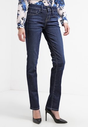 Kvinde iført mørkeblå jeans med lige ben, sorte højhælede sko og en hvid bluse med blå blomsterprint, stående mod en hvid baggrund.