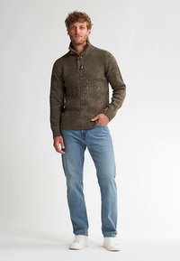 Sweter dziergany w ziemistym zielonym kolorze z wysokim kołnierzem, teksturowanym wzorem i guzikową ozdobą. Połączony z jasnoniebieskimi dżinsami i białymi tenisówkami.