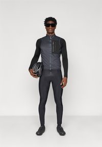 Rattasport, mis sisaldab musta vestiga, millel on lukuga esikülg, sobitatud pikkade varrukatega ja mustade polsterdatud Retuusidega; mudel hoiab käes musta kiivrit.