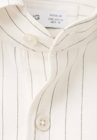 Camisa color crema con sutiles rayas negras verticales. Presenta un cierre de botones y una tela suave y texturizada. Diseño de cuello notable.