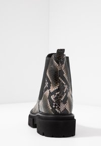 Botín de tobillo con estampado de piel de serpiente en negro y gris, suela gruesa negra, paneles laterales elásticos y tirador, mostrado desde un ángulo trasero sobre una superficie blanca.