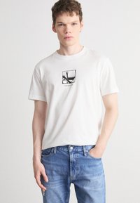 Camiseta de algodón blanca con un gráfico cuadrado de diseño negro, combinada con jeans de mezclilla azul claro. Mangas cortas y cuello redondo.