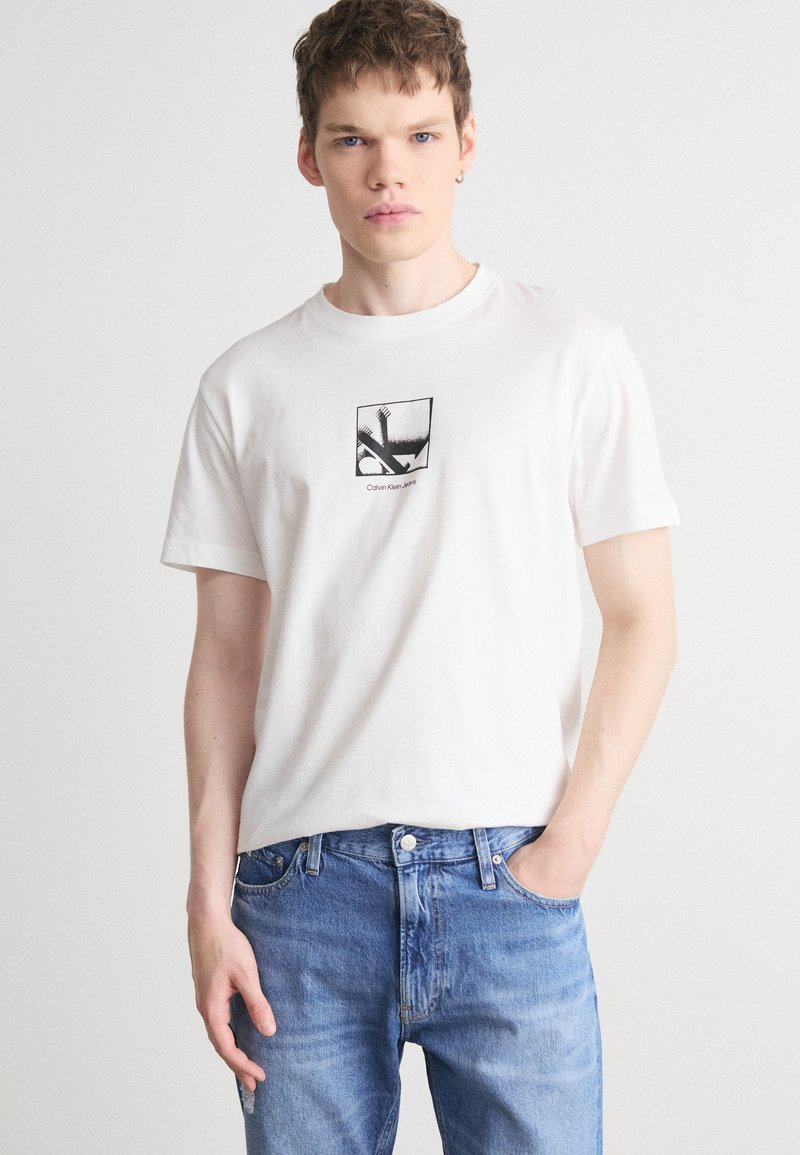 Camiseta de algodón blanca con un gráfico cuadrado de diseño negro, combinada con jeans de mezclilla azul claro. Mangas cortas y cuello redondo.