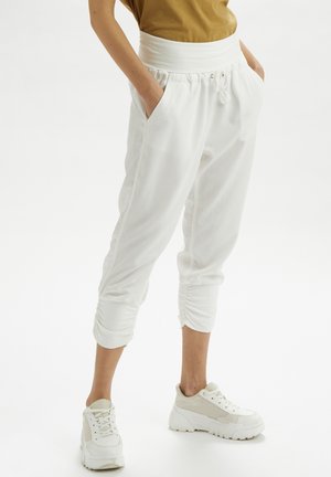 Persona con joggers blancos recortados con puños fruncidos, manos en los bolsillos, camiseta beige y zapatillas blancas gruesas sobre un fondo blanco.