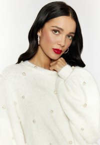Weißer Strickpullover mit lockerer Passform, strukturiertem Stoff und Verzierungen aus kleinen Perlen. Model trägt große Perlenohrringe.