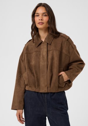 Femme aux longs cheveux foncés portant une veste en daim marron et un jean bleu foncé, debout avec une main dans la poche devant un fond uni.