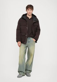 Giacca a vento nera con molte tasche frontali, foderata in nero, abbinata a jeans taper blu chiaro e sneakers grigie. Il modello sta in piedi con le mani nelle tasche.