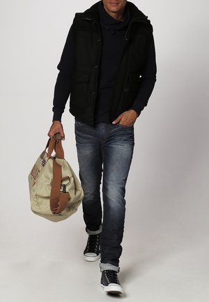 Veste noire sans manches par-dessus un sweat à capuche bleu marine, jean bleu et baskets montantes noires ; portant un sac de voyage beige avec des sangles marron.