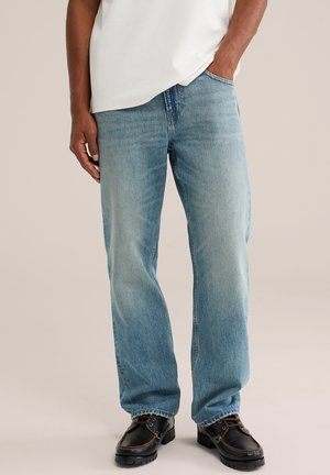 Uomo che indossa jeans azzurri a gamba dritta, scarpe nere di pelle stringate e una camicia bianca con una mano in tasca.