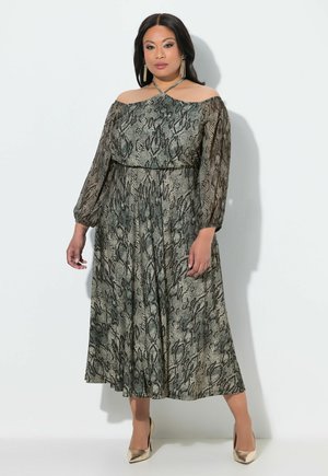 Frau steht, trägt ein schulterfreies, langärmeliges Kleid mit Schlangenprint und goldene High Heels vor einfachem hellem Hintergrund.