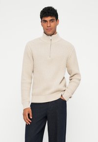 Beige strikket genser med høy krage og halv-glidelås, med et teksturert mønster og ribbestrikkede mansjetter, kombinert med mørke bukser.