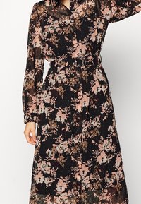 Långärmad svart klänning med rosa och bruna blommönster, knäppning fram, och matchande bälte med fyrkantig spänne vid midjan.