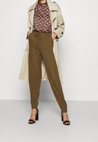 Pantalon de jogging vert olive avec taille à cordon, assorti à un chemisier foncé à motifs, superposé sous un trench-coat beige, et sandales à talons hauts noires.
