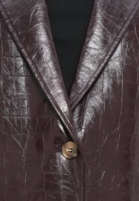 Blazer iz umetnega usnja v burgundski barvi s kroko vzorcem in sijočo površino, ki ima notch ovratnik in enojno zapiranje z rjavim gumbom.