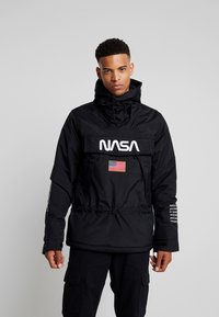 Zwarte jas met een opvallend wit "NASA" logo, Amerikaanse vlag patch, trekkoord onderaan en witte tekst op de mouw. Waterdicht materiaal.