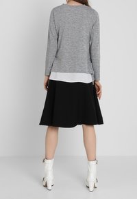 Szara bluzka z długim rękawem nałożona na białą koszulę, w połączeniu z czarną spódnicą w kształcie dzwonu. Modelka nosi białe botki na obcasie.