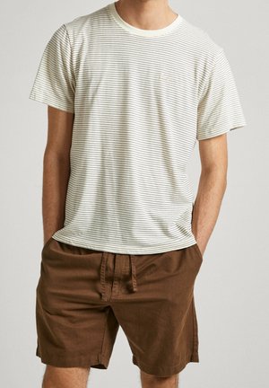 Man draagt een wit t-shirt met korte mouwen met dunne zwarte strepen en bruine short met trekkoord, staand met handen in de zakken.