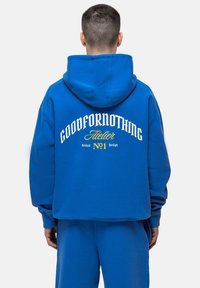 Felpa in cotone blu con una vestibilità ampia. Presenta un grande testo bianco e giallo sul retro. Il testo include "GOODFORNOTHING" e "Atelier."