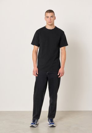 Svart t-shirt i bomull med korta ärmar, kombinerad med löst sittande svarta jeans och sneakers med blå accenter, stående på en ljus betongyta.