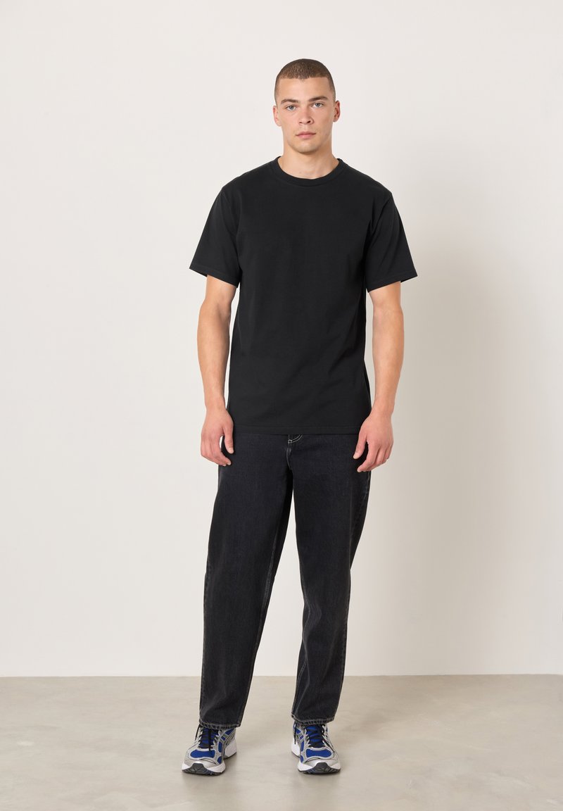 Schwarzes Baumwoll-T-Shirt mit kurzen Ärmeln, kombiniert mit locker sitzenden schwarzen Jeans und blau akzentuierten Turnschuhen, stehend auf einer hellen Betonfläche.