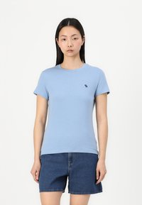 Svetlo modra bombažna majica s kratkimi rokavi, okroglo izrezom in majhnim temno modrim logotipom na prsih. Obuta z jeans kratkimi hlačami.