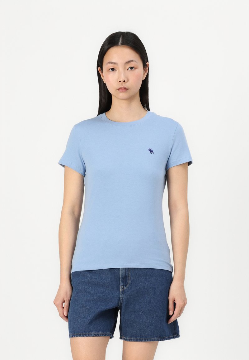 Lyseblå bomulls t-skjorte med korte ermer, rund hals og en liten mørkeblå logo på brystet. Brukt med denimshorts.