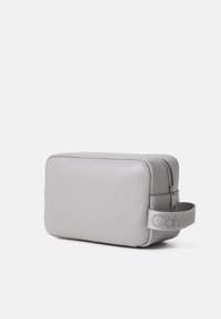 Pochette rectangulaire en cuir gris avec fermeture éclair, sangle en tissu et détail de logo embossé. Texture lisse avec un design minimaliste.