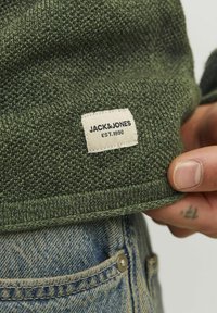 Zöld kötött pulóver texturált mintával, amelynek alsó részén egy "JACK&JONES EST. 1990" feliratú varrott címke található, világoskék farmernadrággal párosítva.