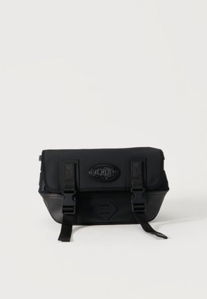 Geantă crossbody Moschino neagră, cu clapetă frontală, două curele cu catarame și patch cu logo pe un fundal simplu, deschis.