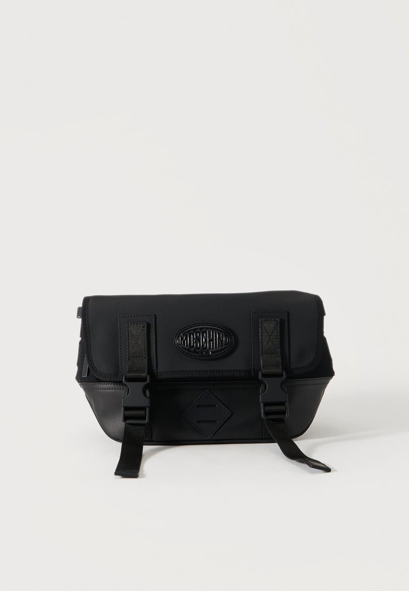 Μαύρο τσαντάκι Moschino χιαστί με μπροστινό καπάκι, δύο λουράκια με αγκράφες και έμβλημα λογότυπου σε απλό ανοιχτό φόντο.