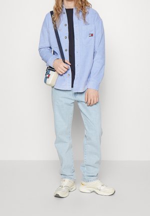 Světlá modrá kordurová košile s knoflíky, tmavě modré tričko, světlé modré džíny a béžovo-šedé tenisky se žlutými detaily. Malá kabelka přes rameno.