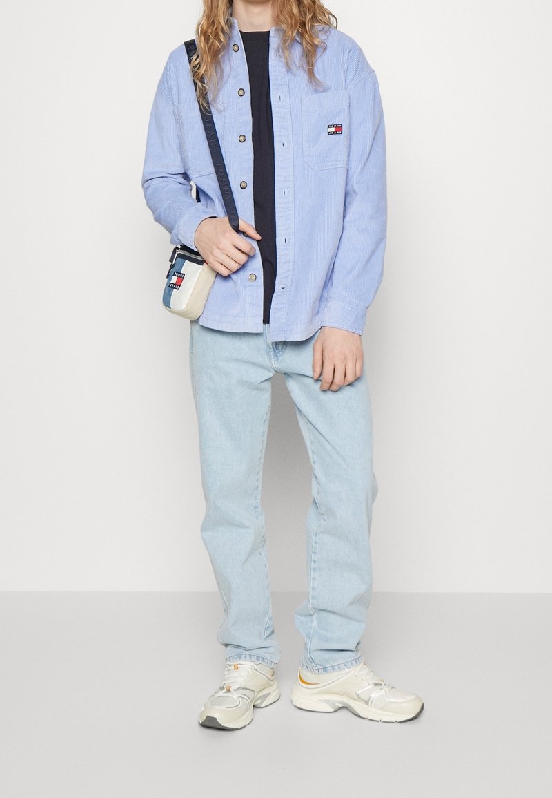 Jasnoniebieska koszula sztruksowa z guzikami, granatowy t-shirt, jasnoniebieskie dżinsy oraz beżowo-szare sneakersy z żółtymi akcentami. Mała torebka na ramię.