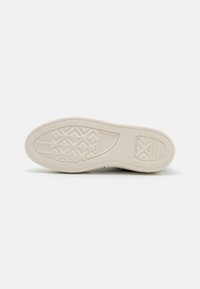 Podeszwa sneakersów, wykonana z teksturowanego, beżowego materiału gumowego z diamentowym wzorem i detalem z logo w pobliżu pięty.