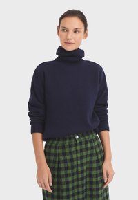 Marinefarbener Rollkragenpullover mit geripptem Halsausschnitt und Bündchen, kombiniert mit einem grün-schwarz karierten Rock, der mit einem knöpfbaren Detail auf der Vorderseite versehen ist.