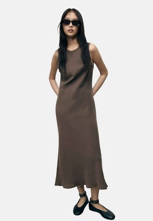 Robe maxi sans manches de couleur marron avec une légère forme en A, texture lisse et couleur unie. Portée avec des ballerines noires et des lunettes de soleil.
