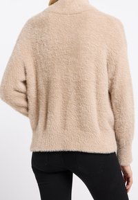 Beige konstpälsjumpa med hög krage, avslappnad passform och texturerad yta, kombinerad med svarta tighta byxor.