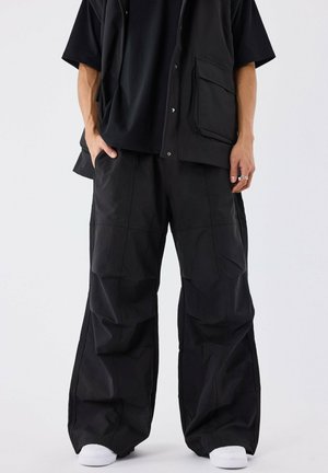 Pantalons noirs surdimensionnés en tissu texturé, avec des jambes larges, des détails de couture et une coupe décontractée, associés à un haut noir.