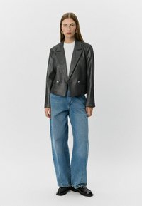 Glanzend zwarte cropped blazer met revers en twee knopen aan de voorkant, gecombineerd met losse blauwe jeans en zwarte schoenen, tegen een lichte achtergrond.