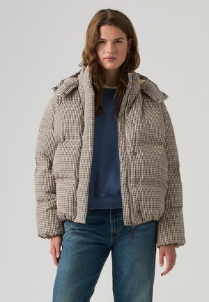 Jeune femme aux cheveux bruns ondulés portant une veste matelassée beige à carreaux, un sweat-shirt bleu et un jean bleu sur un fond clair uni.