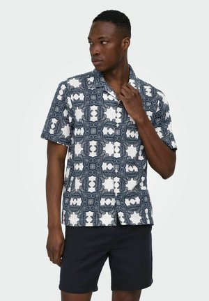 Homme portant une chemise à manches courtes boutonnée avec des motifs ornés bleus et blancs et un short foncé, regardant vers sa gauche.