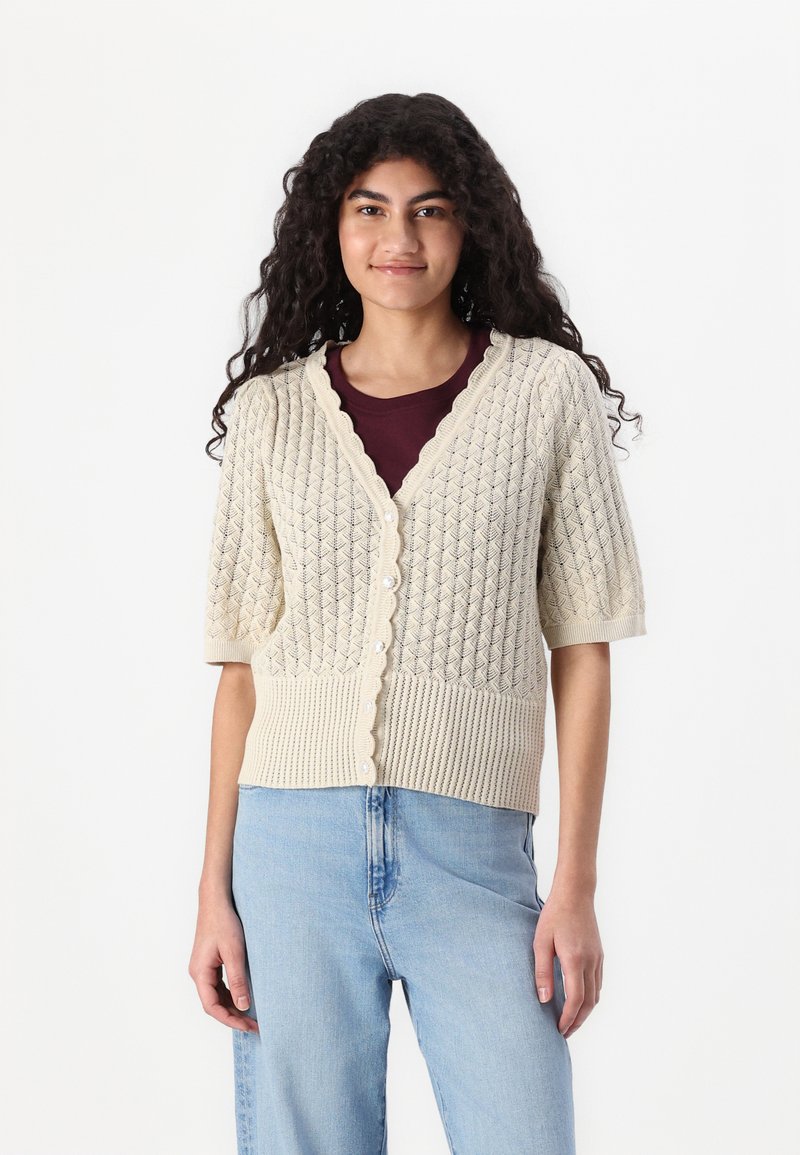 Short-sleeve cardigan in cream featuring a textured diamond pattern, scalloped neckline, and button front, paired with a maroon top.