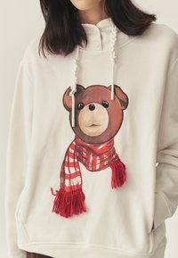 Biela mikina s potlačou medveďa nosiaceho červeno-biely pruhovaný šál. Má okrúhly výstrih a kapucňu so sťahovacou šnúrkou.
