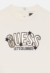 Sweatshirt blanc avec "GUESS" en texte métallisé doré et noir, et "LET'S CELEBRATE" en dessous, accompagné d'étoiles noires qui accentuent le design.