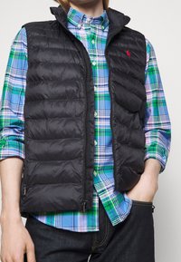 Osoba v černé prošívané vestě přes modrou, zelenou a bílou kostkovanou košili s jednou rukou v kapse tmavých džínů.