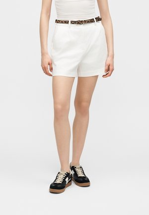 Personne portant un short blanc taille haute avec une ceinture léopard, un haut blanc et des baskets noires avec des lacets blancs, debout sur un fond uni.