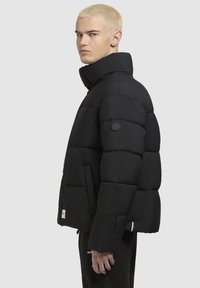 Junge Person mit kurzen blonden Haaren, die eine schwarze Steppjacke mit hohem Kragen und schwarze Hosen vor einem schlichten hellen Hintergrund trägt.