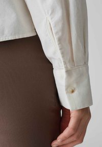 White corduroy shirt cuff with a single button closure. The cuff features a slit detail and is paired with a brown garment.