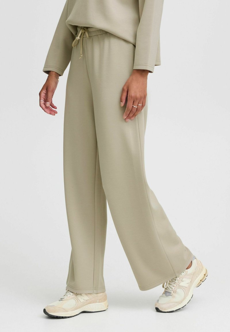Pantalones de chándal color beige claro con un corte relajado, cintura con cordón y costuras laterales. Combinados con zapatillas deportivas en beige y gris.