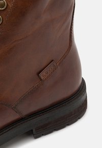 Braune Lederstiefel mit einer strukturierten Oberfläche, genähten Akzenten und einer Gummisohle mit einem robusten Profil. Die Markenkennzeichnung ist an der Seite sichtbar.