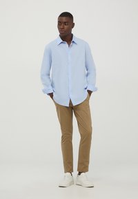 Camicia azzurra chiara con maniche lunghe, abbinata a pantaloni beige e sneakers bianche, in piedi su uno sfondo semplice.